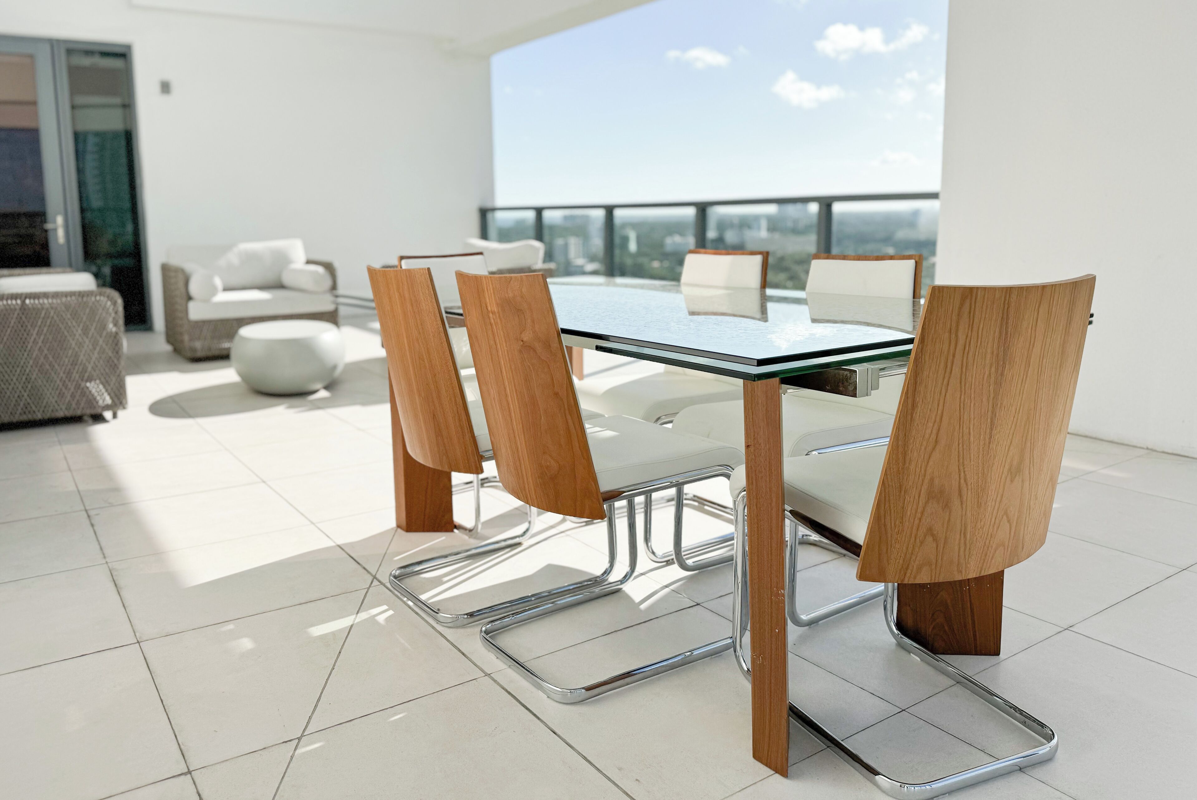 A modern dining area with a glass table and wooden chairs, a white sofa, and a city view.