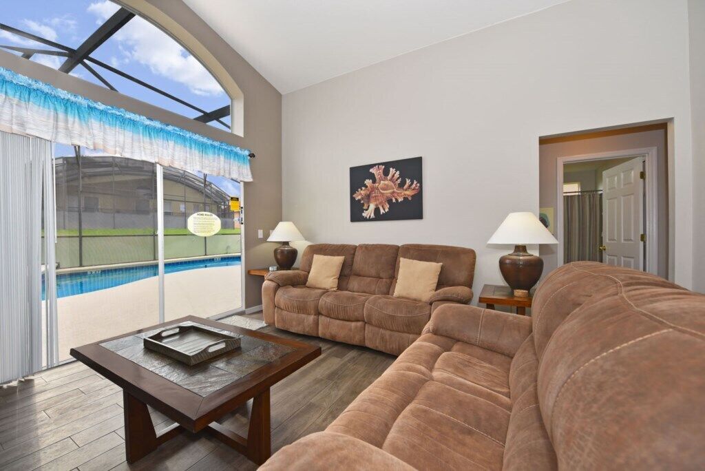 A living room with a large window overlooking a pool, a brown sofa, a coffee table, and a framed picture on the wall.