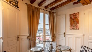 A room with a dining table set for two, a window with curtains, a balcony with a railing, and a painting on the wall. - Key West - charming studio ON the port of Honfleur - Superb 360° view (Honfleur)