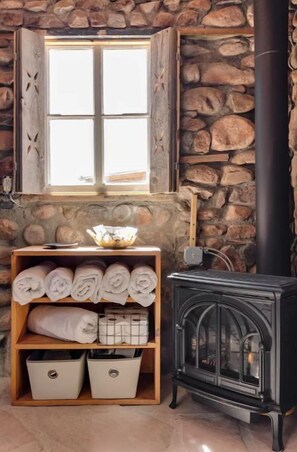 A stone wall, a window with curtains, a wooden shelf with towels, and a black wood-burning stove. - La Biblioteca: Eclectic Cottage in OKeeffe Country (Abiquiu)