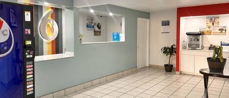 Un vestíbulo de hotel con una máquina expendedora, una recepción, una cafetera y una planta en maceta.