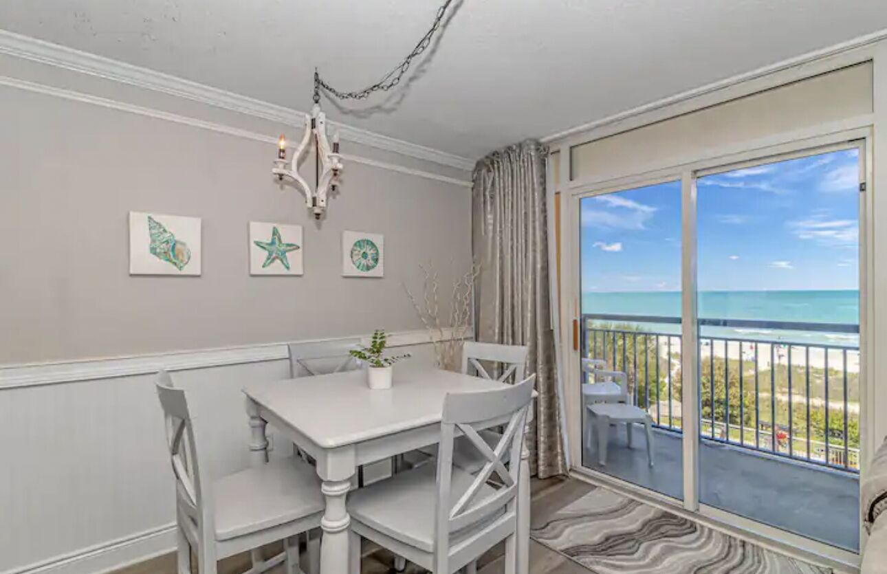 A dining area with a white table and chairs, a chandelier, and a view of the ocean through a sliding glass door.