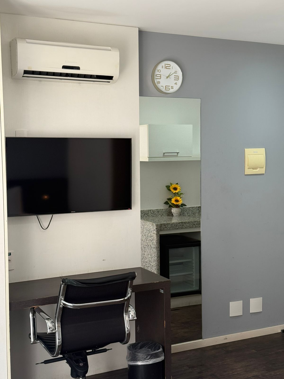 A modern living room with a flat-screen TV, a wall-mounted clock, a small table with a chair, and a decorative vase with sunflowers.
