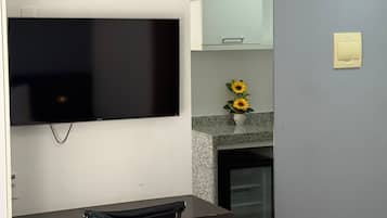 A modern living room with a flat-screen TV, a wall-mounted clock, a small table with a chair, and a decorative vase with sunflowers.