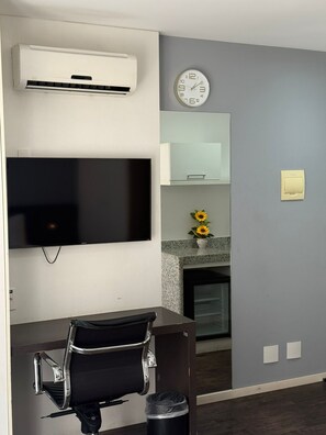 A modern living room with a flat-screen TV, a wall-mounted clock, a small table with a chair, and a decorative vase with sunflowers. - Suites Pampulha Hotel (Belo Horizonte)