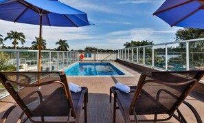 Un espace aménagé au bord de la piscine, comprenant deux chaises longues, un parasol bleu et une rambarde en verre donnant sur la piscine.