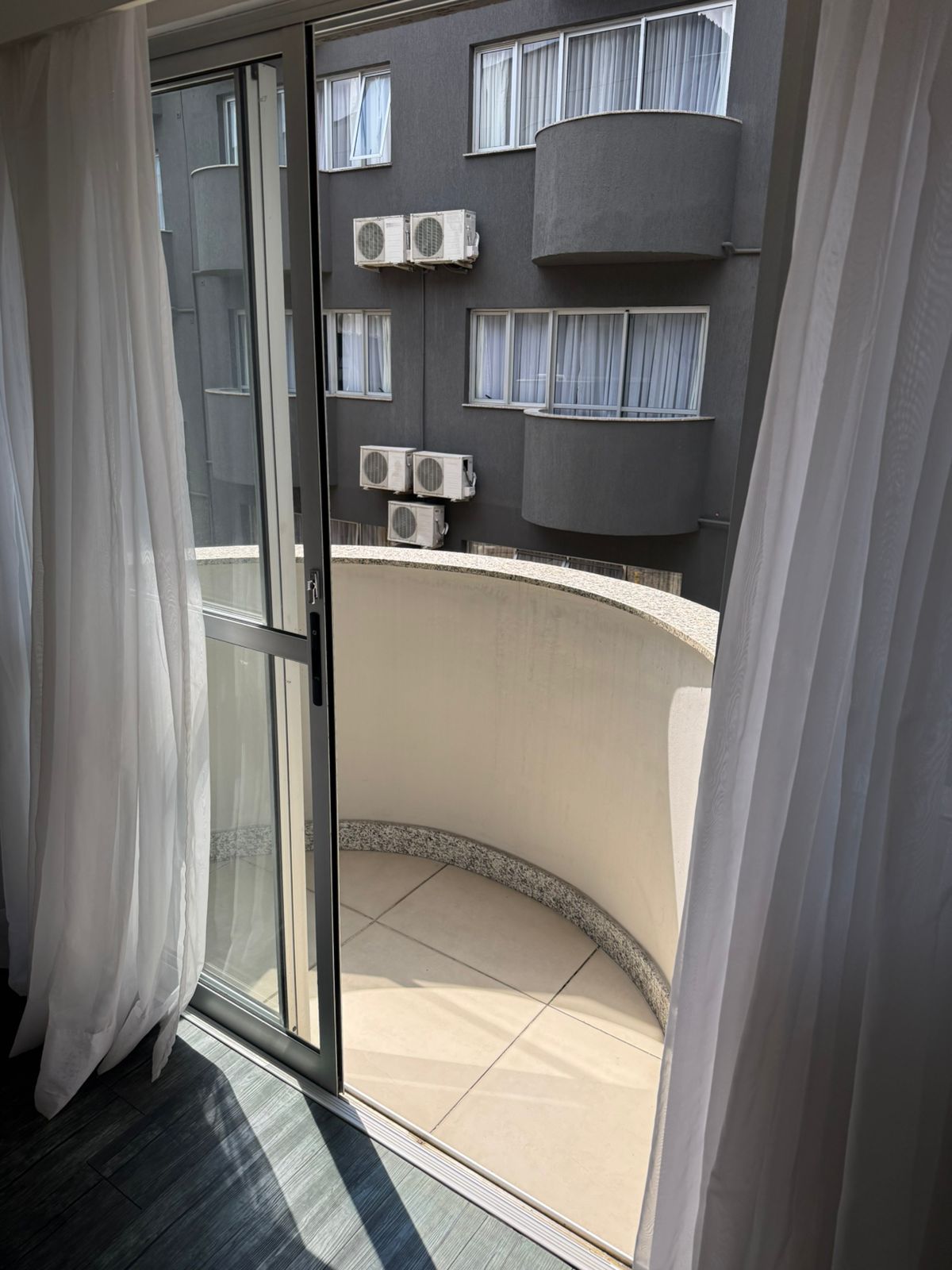 Une pièce dotée d’une porte-fenêtre coulissante donnant sur un balcon équipé de climatiseurs et offrant une vue sur un immeuble à multiples fenêtres.