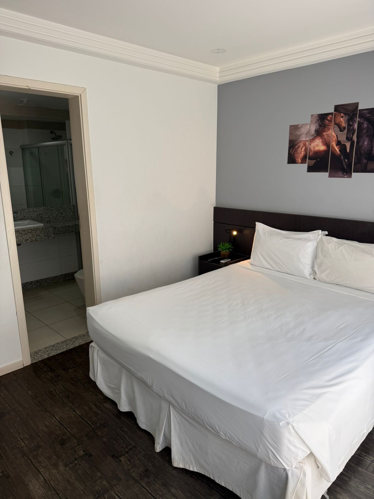 Un dormitorio con una cama grande, una mesita de noche, una obra de arte en la pared y un baño visible a través de una puerta abierta.