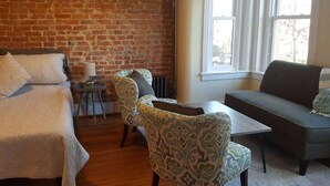 Una habitación con pared de ladrillo, una cama, un sofá, dos sillones, una mesita y una ventana con persianas.