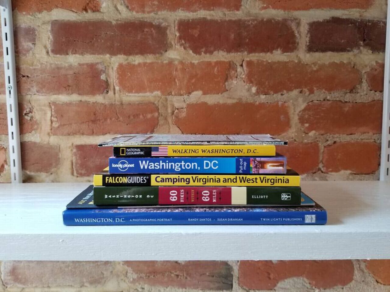 Une pile de livres sur une étagère blanche, adossée à un mur de briques.