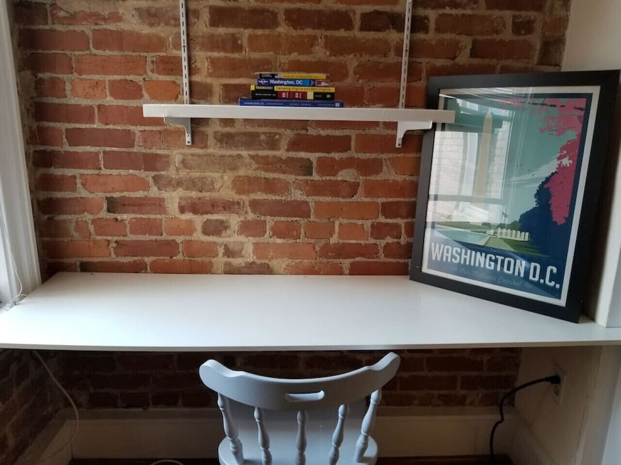 A white desk with a white chair in front, a brick wall, and a framed map of Washington D.C. on the wall.