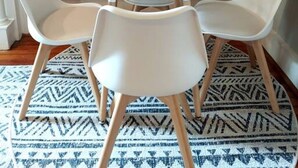 A round white table with four white chairs, set on a patterned rug, against a brick wall.