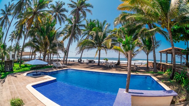 A swimming pool with a tiled deck, surrounded by palm trees and a beach view.