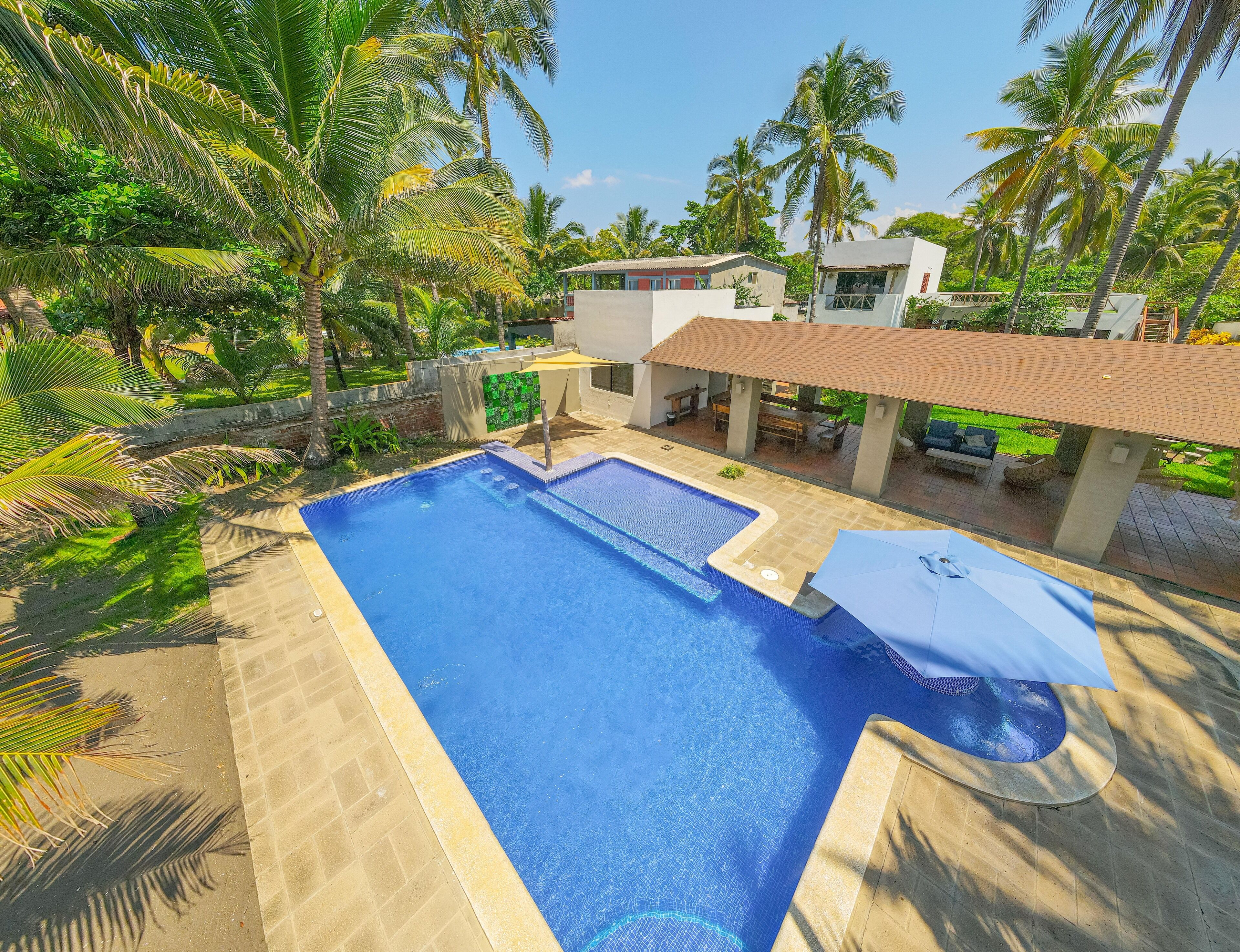 A rectangular swimming pool with a tiled deck, surrounded by palm trees and a covered patio area with outdoor furniture.