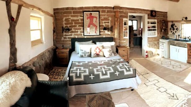 A rustic bedroom with exposed brick walls, a bed with patterned bedding, a wooden ceiling, and a view into the kitchen area. - Luscious Adobe Studio in Gorgeous Abiquiu! (Abiquiu)
