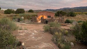 A rustic cabin with a stone foundation and a small wooden structure, surrounded by desert vegetation and mountains in the background. - Luscious Adobe Studio in Gorgeous Abiquiu! (Abiquiu)