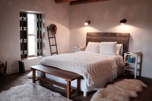 A bedroom with a wooden headboard, a white bedspread, a bench, a small bedside table, and a nightstand. - Casa Tocaya: Adobe Gem in O'Keeffe country! (Abiquiu)
