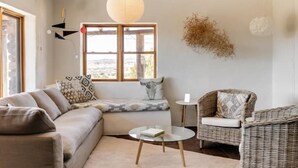 A cozy living room with a sofa, a round glass coffee table, a wicker chair, and a large window with a view. - Casa Tocaya: Adobe Gem in O'Keeffe country! (Abiquiu)