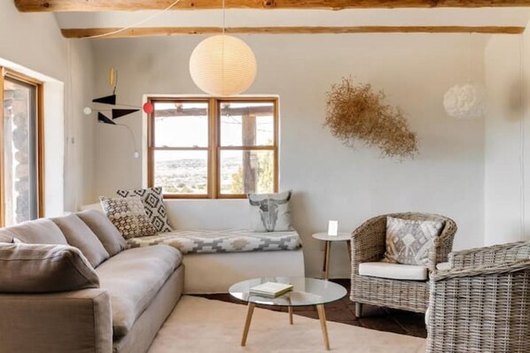 A cozy living room with a sofa, a round glass coffee table, a wicker chair, and a large window with a view. - Casa Tocaya: Adobe Gem in O'Keeffe country! (Abiquiu)