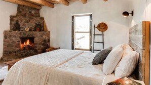 A cozy bedroom with a stone fireplace, wooden beams, a bed with pillows, and a ladder against the wall. - Casa Tocaya: Adobe Gem in O'Keeffe country! (Abiquiu)