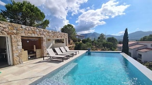 Une piscine avec des chaises longues, un bâtiment en pierre et un paysage montagneux en arrière-plan.