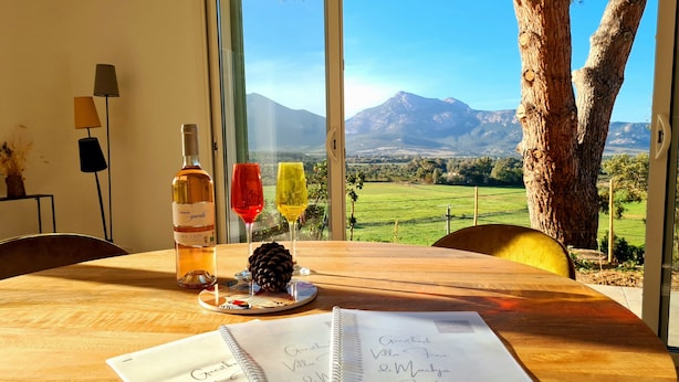 Une table avec une bouteille de vin, deux verres, une pomme de pin et un carnet contenant des notes manuscrites.
