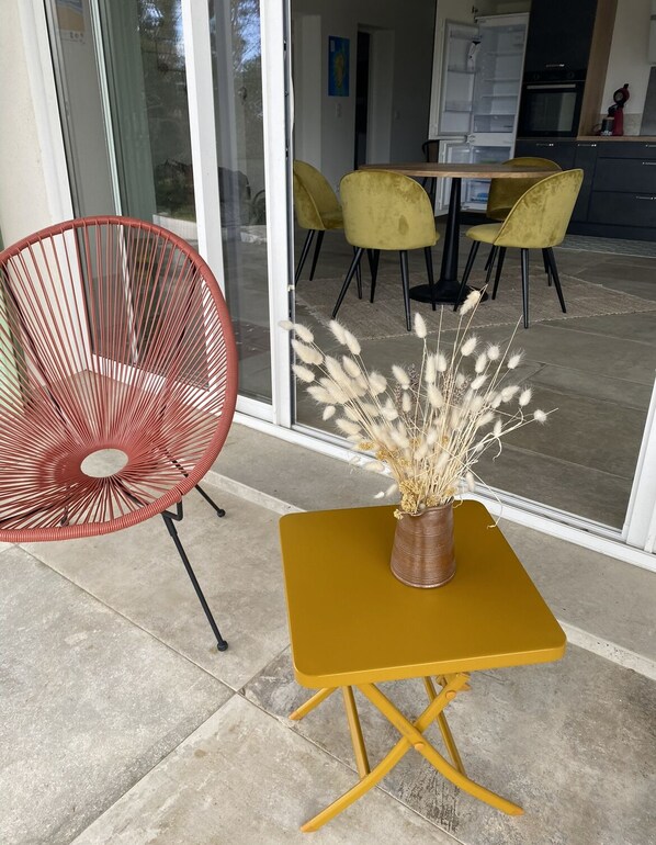 Un espace extérieur moderne, aménagé avec des sièges, comprenant une chaise rouge, une table jaune et un vase rempli de plumes d’herbe des pampas.
