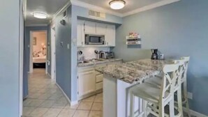 A compact kitchen with white cabinets, a microwave, and a small island with bar stools. - Coastal Condo - Beach view, resort (Galveston)