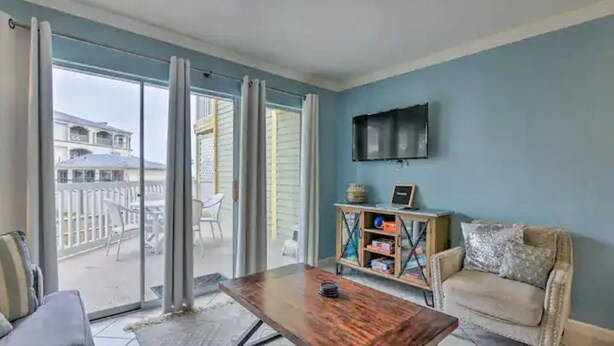 A living room with a wooden coffee table, a sofa, a TV mounted on the wall, and a sliding glass door leading to an outdoor area with chairs and a table. - Coastal Condo - Beach view, resort (Galveston)