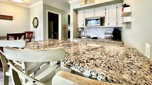 A kitchen with granite countertops, white cabinets, and a dining area with wooden chairs. - Coastal Condo - Beach view, resort (Galveston)