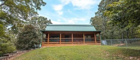 A wooden cabin with a green roof, surrounded by trees and a grassy area.