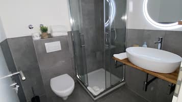 Un baño moderno con ducha envuelta en vidrio, inodoro blanco y un espejo redondo grande sobre un lavamanos blanco.