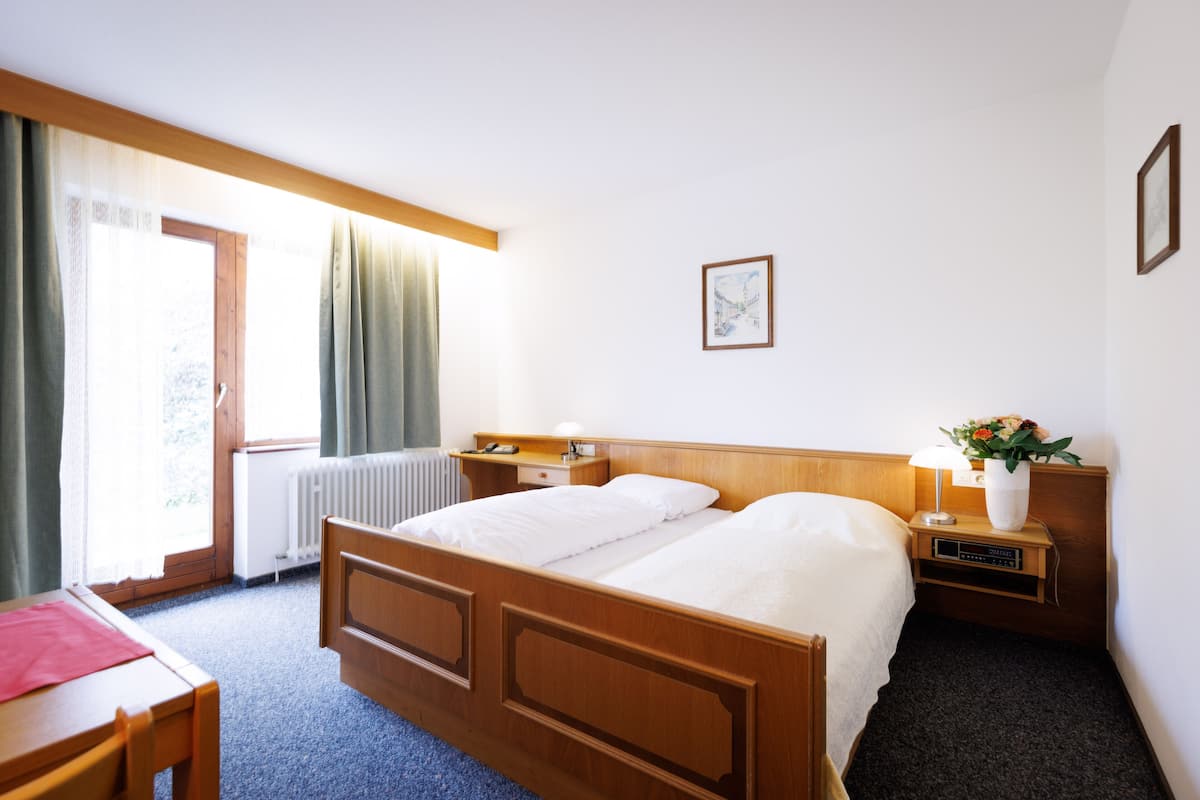 Hotelkamer met een houten bed, een bureau, een stoel, een nachtkastje met lamp en een raam met gordijnen.