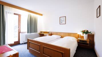 Une chambre d’hôtel avec un lit en bois, un bureau, une chaise, une table de chevet avec une lampe et une fenêtre avec des rideaux.
