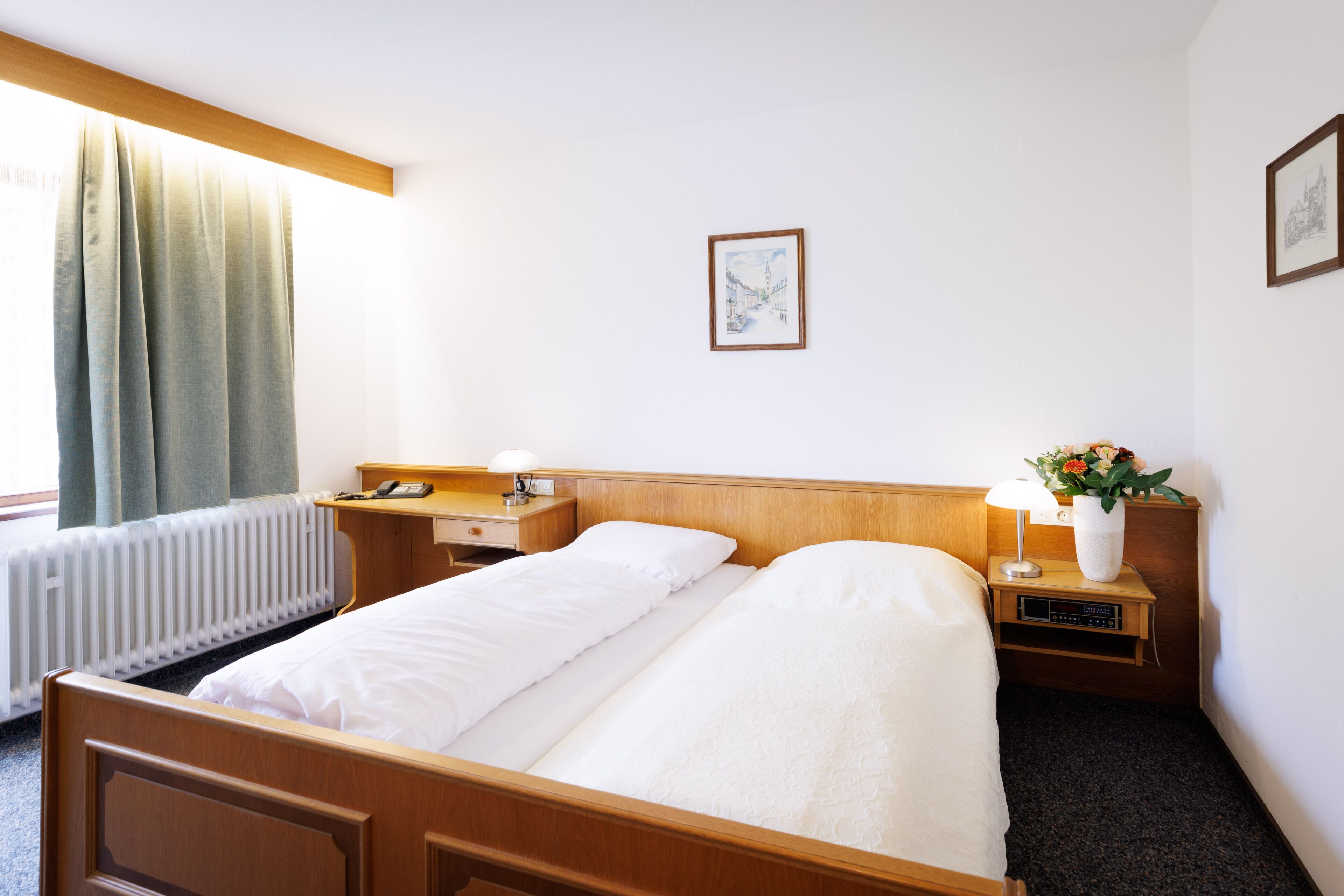 A hotel room with a wooden bed, a bedside table with a lamp, a small nightstand with a vase of flowers, and a framed picture on the wall.
