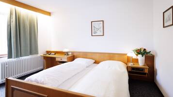 A hotel room with a wooden bed, a bedside table with a lamp, a small nightstand with a vase of flowers, and a framed picture on the wall.