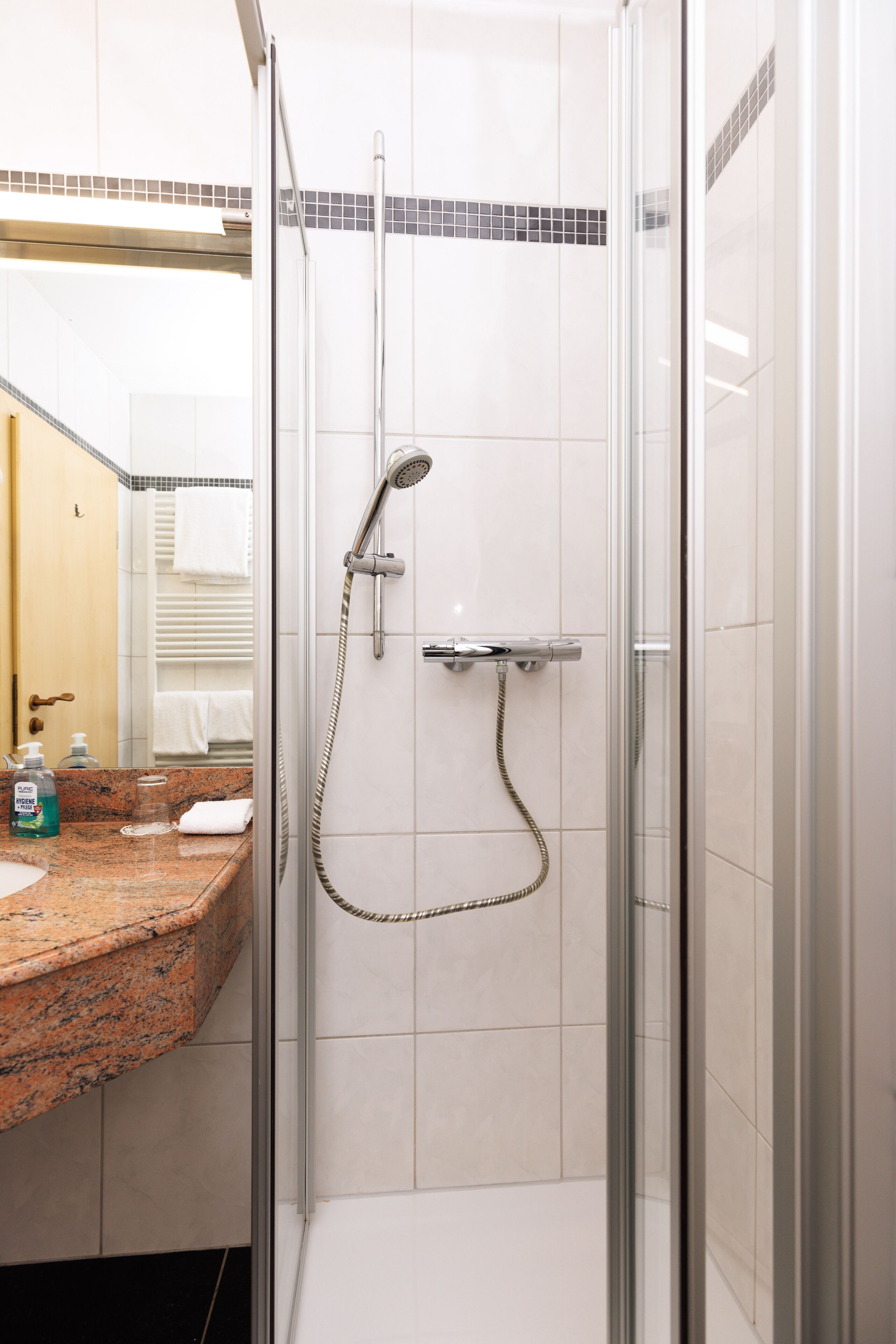 A shower with a glass door, a tiled wall, and a granite countertop with a sink and toiletries.
