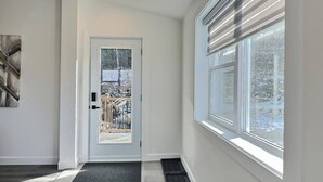 A modern hallway with a glass door leading outside, a window with blinds, and a black mat at the entrance.