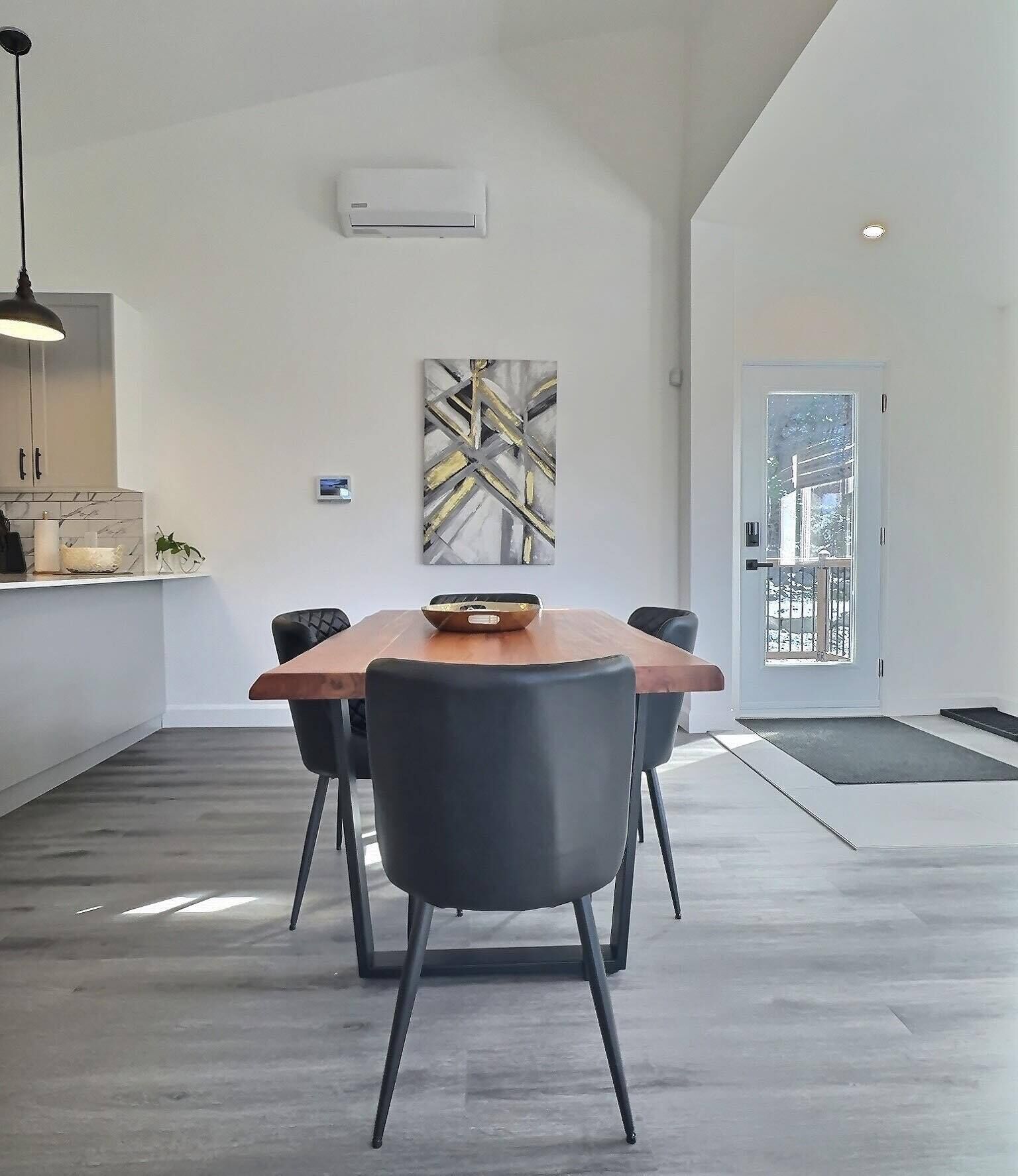 Ein zeitgemäßer Essbereich mit Holztisch und schwarzen Stühlen, eine Küche mit weißen Schränken und ein großes abstraktes Gemälde an der Wand.