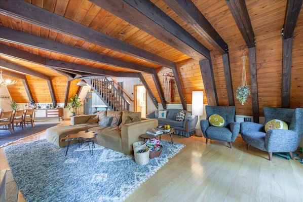 A spacious loft with exposed wooden beams, a staircase, a sofa, armchairs, and a dining area.