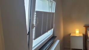 Ein Schlafzimmer mit Fenster, Heizkörper, Nachttisch mit Lampe und Bett mit Kissen.