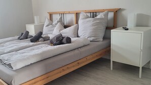 Ein ordentlich bezogenes Bett mit grauen Bettwäsche und zusammengerollten Handtüchern, ein Holzkopfschild, weiße Nachttische und eine Fernbedienung auf einem der Tische.