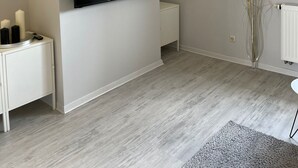 Ein modernes Wohnzimmer mit einem an einer weißen Wand montierten Flachbildfernseher, einem kleinen weißen Konsolentisch mit Kerzen, einer Heizung und einer Vase mit dekorativen Federn.