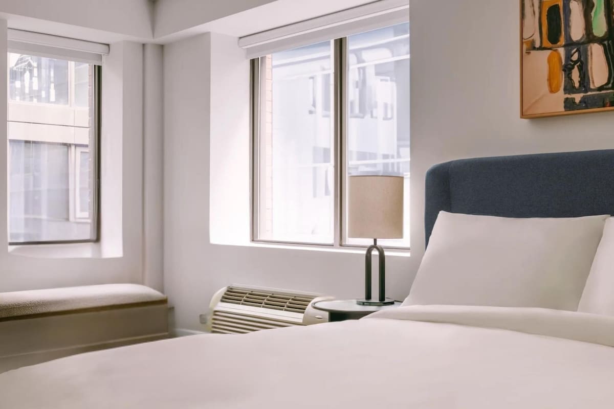 A hotel room with a large bed, a bedside table with a lamp, a heater, and a window with blinds.