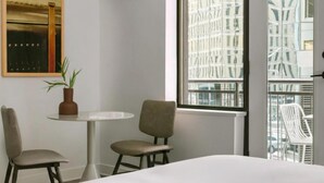 Une chambre d’hôtel avec un lit, deux chaises, une petite table, une plante en pot et une grande fenêtre donnant sur des immeubles.