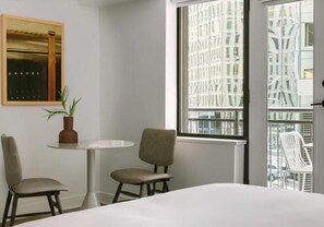 Una camera d'albergo con un letto, due poltrone, un tavolino, una pianta in vaso e un'ampia finestra che si affaccia su edifici.