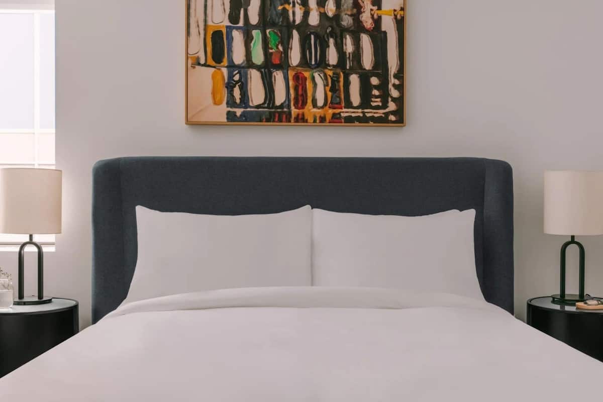 Una cama con cabecera oscura, ropa de cama blanca y un cuadro abstracto colorido en la pared.