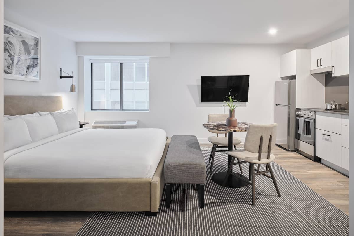 Una habitación de hotel moderna con una cama grande, un área de comedor con mesa y sillas, una cocina con electrodomésticos y un televisor de pantalla plana montado en la pared.