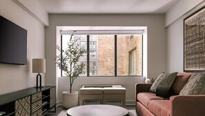 Un salon moderne avec un canapé, une table basse, un téléviseur et une fenêtre donnant sur un immeuble en briques.
