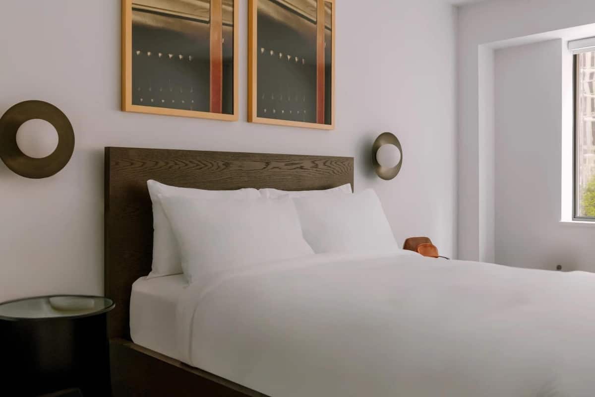 Una cama bien tendida con ropa blanca, cabecera de madera y dos cuadros enmarcados en la pared.